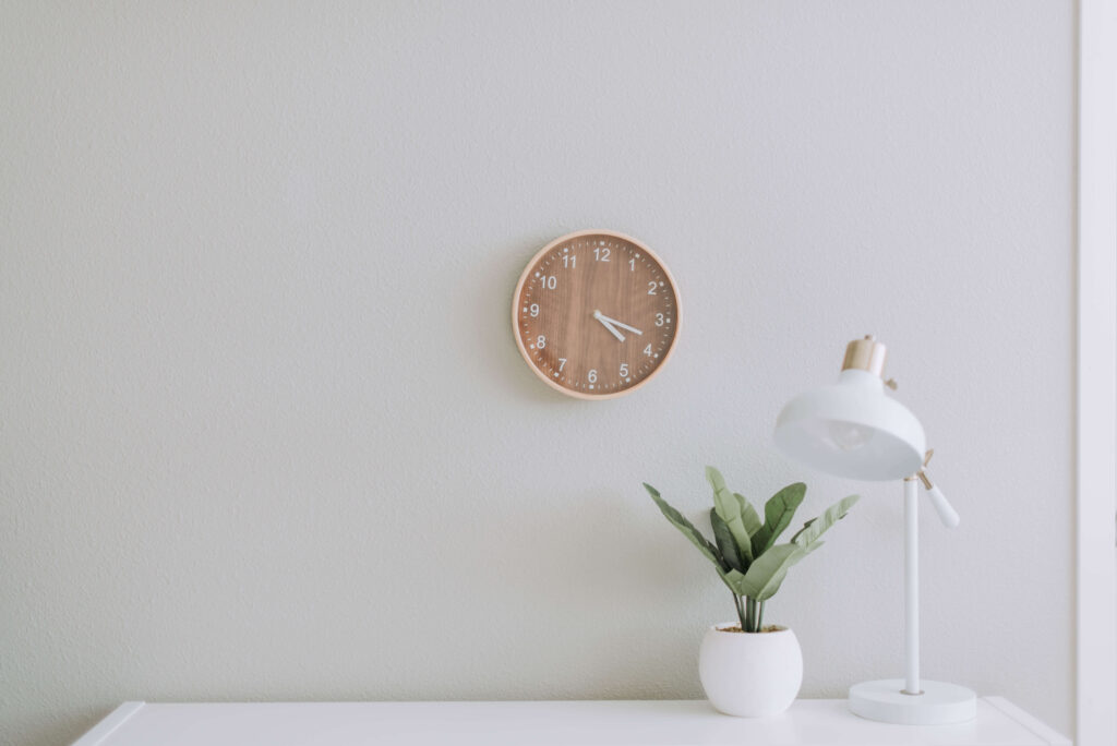 A brown clock is on the wall and a desk plant and lamp rest on a white desk in an empty room. Sojourner Counseling provides a comfy space for those looking to work with a christian counselor in Raleigh NC. Call today!